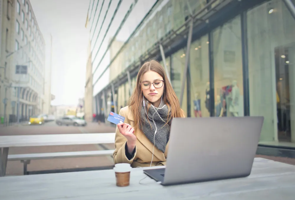 Eine Frau sitzt draußen an Ihrem Laptop und hat eine Kreditkarte in der Hand